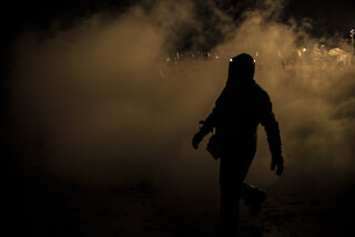 Boris, a water protector, walks between fires at the front lines carrying logs with embers on them from fire to fire as they are pummeled by police fire hoses.